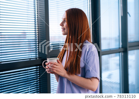 Happy redhead young woman drinking coffee and watching on sunset through the window after working day. 78313110
