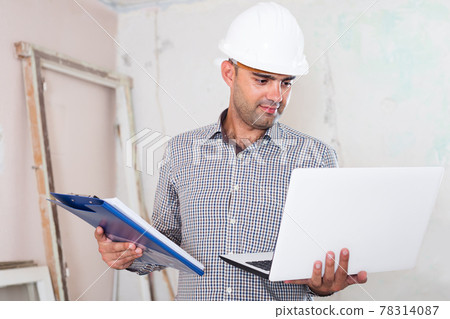 Portrait of positive architect working at the notebook in helmet with documents 78314087