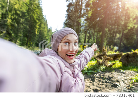 Young adult caucasian beautiful happy smiling woman making selfie shot by cell phone pointing finger backward during walk in scenic green woods pine forest river. Person enjoy walking nature outdoors 78315401