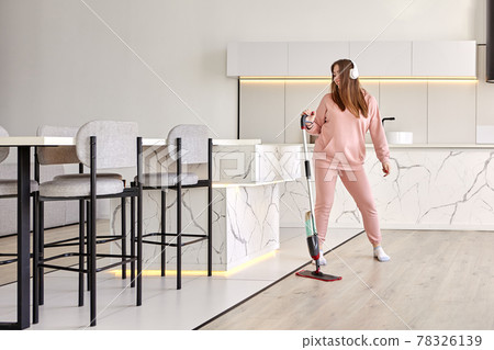 Young Millennial woman wipes floor in kitchen, listening to music through wireless headphones and dancing. 78326139