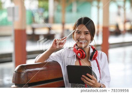 Cheerful young woman traveller in headphone holding digital tablet and smiling to camera while sitting in train station. 78339956
