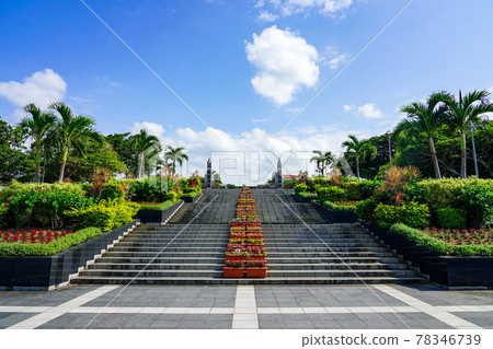 A stone staircase with a beautiful flower bed leading to the blue sky and blue sea of Okinawa and the central gate of Kaiyohaku Park 78346739