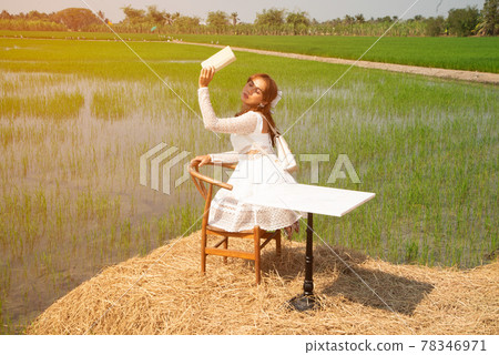 Beautiful Asian woman sitting on a table in a rice field holding a book and stretching it to block sunlight. 78346971