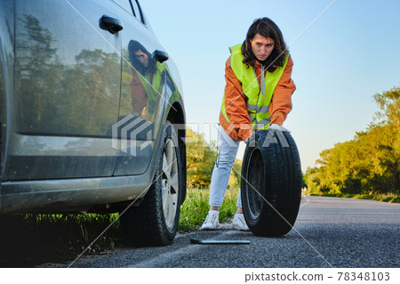 The girl changes the damaged wheel of the car. Green vest. Spare wheel. Traffic accident. Travel by car. 78348103