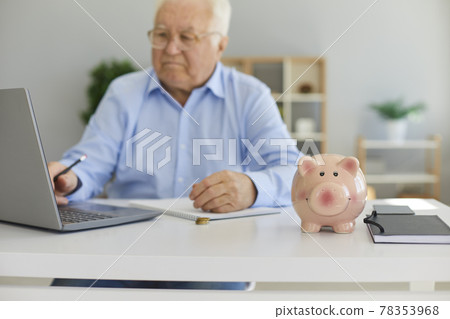 Portrait of a senior man working on a laptop in a bright office with a piggy bank in the foreground. Portrait of a senior man working on a laptop in a bright office with a piggy bank in the foreground. 78353968
