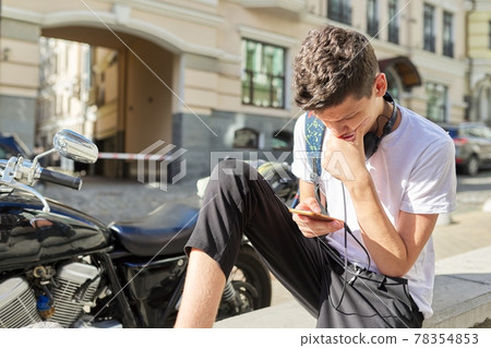 Boy teenager 16,17 years old, using smartphone, sitting on street of summer city 78354853