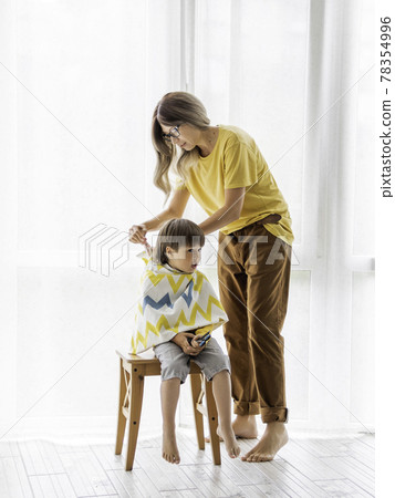 Mother prepares to cut her son's hair by herself. Little boy sits, covered with cloth, and holds pair of scissors. New normal in quarantine of coronavirus COVID-19. Self care at home lifestyle. 78354996
