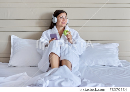 Mature woman relaxing in bed in headphones with smartphone, green apple Mature woman relaxing in bed in headphones with smartphone, green apple 78370584