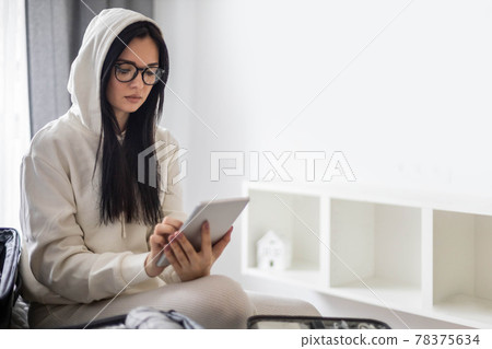 A beautiful young girl in a white casual suit sits on the bed and uses a tablet. 78375634