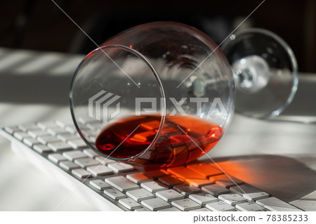 A glass of red wine lies on the keyboard on a white table with a shade from the blinds A glass of red wine lies on the keyboard on a white table with a shade from the blinds 78385233