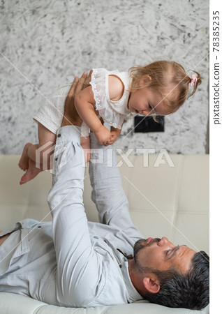 A Caucasian man lies on the couch and holds a little daughter above him. Caring loving dad raised the child up. Happy fatherhood. A Caucasian man lies on the couch and holds a little daughter above him. Caring loving dad raised the child up. Happy fatherhood. 78385235