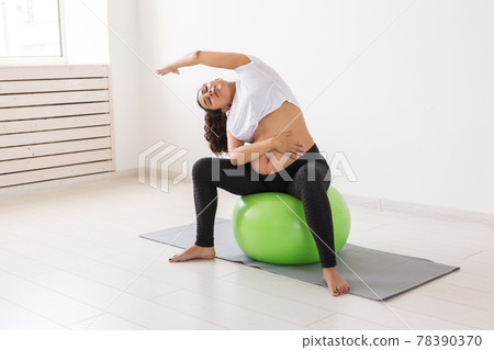 A young pregnant woman doing exercise using a fitness ball while sitting on a mat. A young pregnant woman doing exercise using a fitness ball while sitting on a mat. 78390370