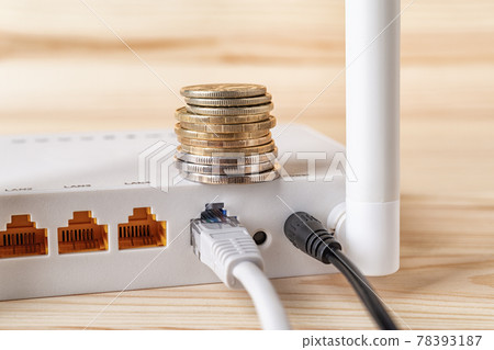 Stack of coins on the white Wi-Fi wireless router with connected cables on a wooden desk. 78393187