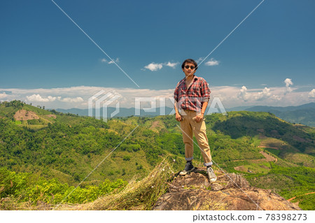Asian man standing on a rock against a backdrop of mountains and sky. A 35-year-old man wearing a striped shirt and brown trousers with sunglasses stands on a rock. Asian man standing on a rock against a backdrop of mountains and sky. A 35-year-old man wearing a striped shirt and brown trousers with sunglasses stands on a rock. 78398273