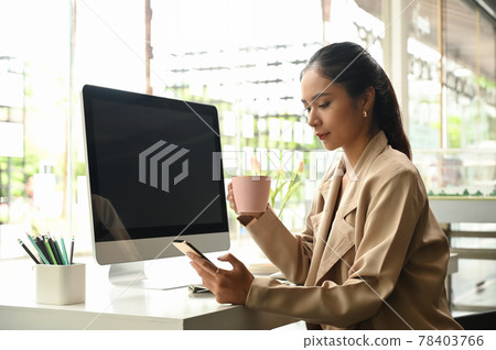 Portrait of young businesswoman looking at her smartphone while drinking coffee in the morning. Portrait of young businesswoman looking at her smartphone while drinking coffee in the morning. 78403766