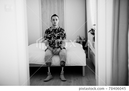 Black and white portrait of disabled young man sitting on bed indoors at home, leg prosthetic concept. 78406045