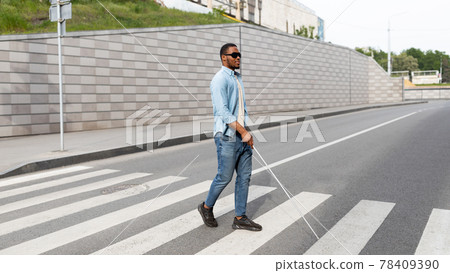 Young black visually impaired man wearing dark glasses, walking across city street, using cane Young black visually impaired man wearing dark glasses, walking across city street, using cane 78409390