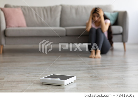 Stressed young Indian woman sitting on floor, grabbing her head in terror, looking at empty scales, feeling overweight 78419102