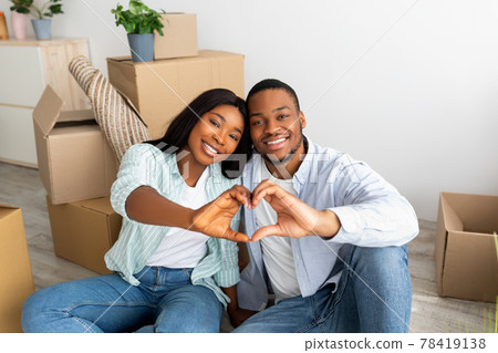 Love nest. Happy black couple sitting among moving boxes, making heart shape gesture with fingers and smiling to camera 78419138