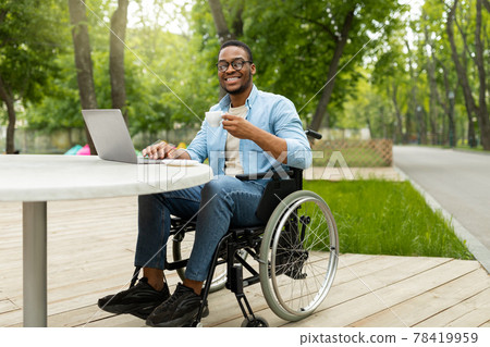 Remote job for disabled people. Joyful black guy in wheelchair working online, using laptop at outdoor cafe 78419959