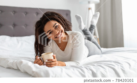 Black woman lying on bed, holding cup of hot coffee 78420101