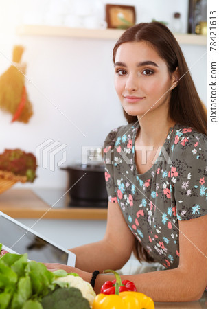 An attractive young dark-haired woman chooses the recipe for a delicious meal while sitting at the table in sunny kitchen An attractive young dark-haired woman chooses the recipe for a delicious meal while sitting at the table in sunny kitchen 78421613