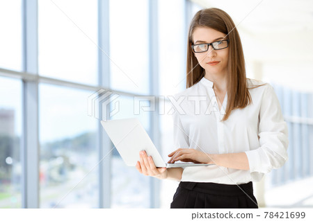 Beautiful female specialist with laptop computer standing in modern office and smiling charmingly. Working on design, data analysis, plan strategy. Business people concept 78421699