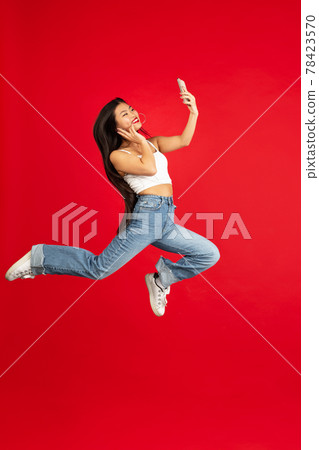 always on mobile. Full length of pretty young woman taking phone while jumping against red studio background. always on mobile. Full length of pretty young woman taking phone while jumping against red studio background. 78423570
