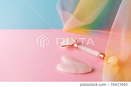 Gua sha and quartz facial roller for skincare massage on blue pink background with colorful rainbow organza drapery 78423992