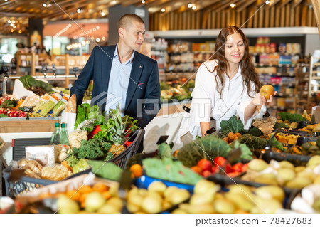 Positive young couple with a grocery cart in the supermarket, choose pears 78427683