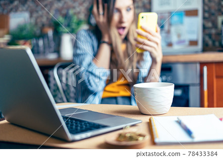 Distance learning online education and work. Business woman having a facetime video call. Surprised girl working from home office kithcen and drinking coffee. Using computer and cellphone. 78433384