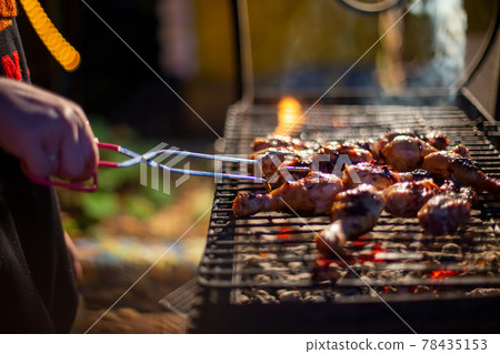 a human hand turns chicken drumsticks on a barbecue grill with grilling tongs. cooking food on an open fire in the evening. backyard party 78435153