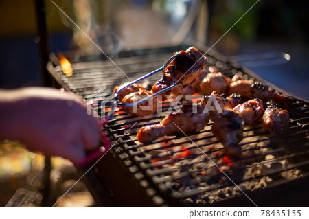 a human hand turns chicken drumsticks on a barbecue grill with grilling tongs. cooking food on an open fire in the evening. backyard party a human hand turns chicken drumsticks on a barbecue grill with grilling tongs. cooking food on an open fire in the evening. backyard party 78435155