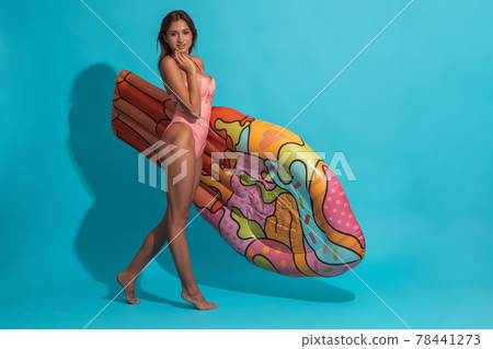Smiling girl in bathing suit carrying swimming mattress on blue background 78441273