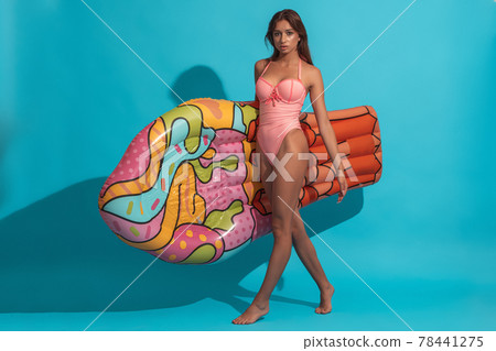Girl in swimsuit walking on blue background with inflatable mattress Girl in swimsuit walking on blue background with inflatable mattress 78441275