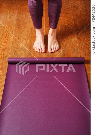 The girl's feet stand in front of an unfolded Yoga mat on the wooden floor. 78441526