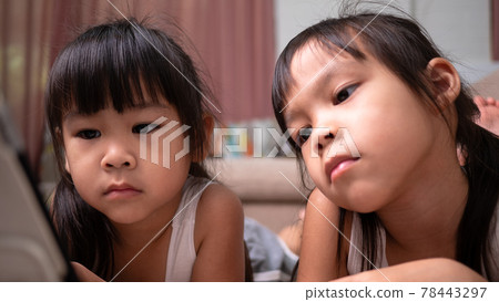 Two sibling girls lying on the sofa and watching digital tablets together in the living room. 78443297