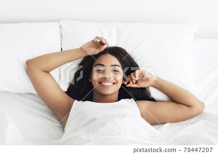 Relaxed black lady waking up and smiling to camera, feeling happy, lying on white bedsheets, top view 78447207