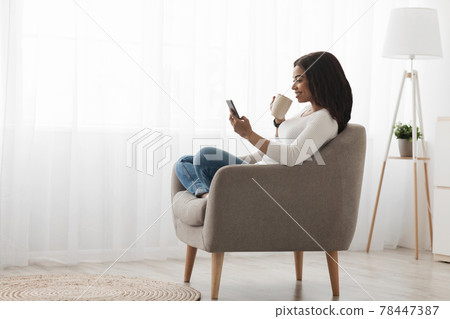Domestic rest. Calm black woman using smartphone, sitting with cup of coffee near window, enjoying morning, free space Domestic rest. Calm black woman using smartphone, sitting with cup of coffee near window, enjoying morning, free space 78447387