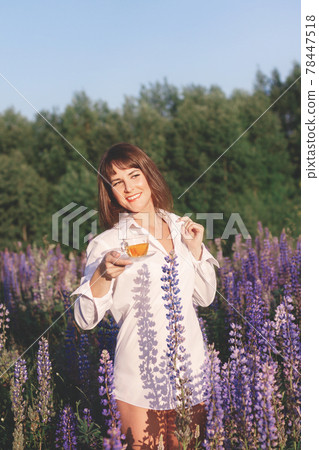 Woman in white shirt in field of lupins and holds glass cup of floral tea in summer Woman in white shirt in field of lupins and holds glass cup of floral tea in summer 78447518