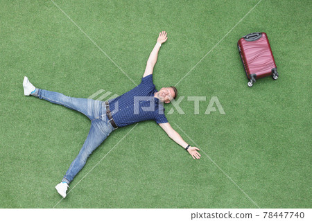 Happy man lies on green lawn with suitcase top view 78447740