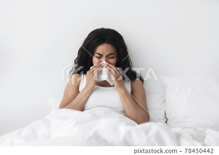 Runny nose, flu and colds. Sick african american lady lying in bed and blowing her nose with napkin Runny nose, flu and colds. Sick african american lady lying in bed and blowing her nose with napkin 78450442
