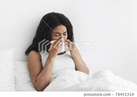 Influenza. Sick african american woman blowing runny nose sneezing, sitting in bed, copy space Influenza. Sick african american woman blowing runny nose sneezing, sitting in bed, copy space 78451319