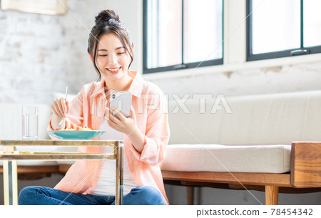 一位年輕女子在客廳吃意大利面時看著她的智能手機 一位年輕女子在客廳吃意大利面時看著她的智能手機 78454342