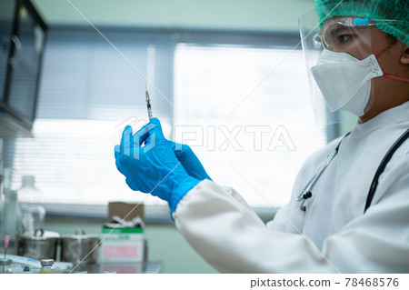 Young doctor in a white uniform wearing a face and face shield prepares a syringe to vaccinate people in the medical center. 78468576