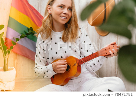 Young millennial hippie woman sitting on balcony play guitar. Music lesson and singer sitting. LGBTQ rainbow flag on background. Online music tuition. Playing instrument and enjoying songs and leisure 78471791