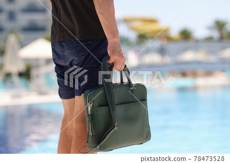 Man tourist in shorts holds business bag in background of hotel pool 78473528