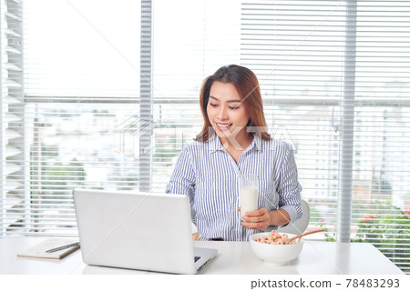 Beautiful young woman sitting at the table in home office, holding glass of milk with cereal, using laptop. 78483293