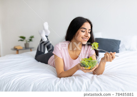 Balanced raw food diet. Lovely Indian woman eating fresh vegetable salad, lying on bed at home with yummy meal Balanced raw food diet. Lovely Indian woman eating fresh vegetable salad, lying on bed at home with yummy meal 78485881