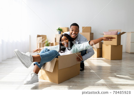 Moving and real estate concept. Joyful black couple having fun and riding in cardboard boxes at new apartment 78485928